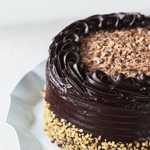 A German Chocolate Cake on a white cake plate with a deeper, more complex flavor profile that balances perfectly, chocolatey layer cakes sandwiched with chocolate fudge and covered in creamy coconut icing.