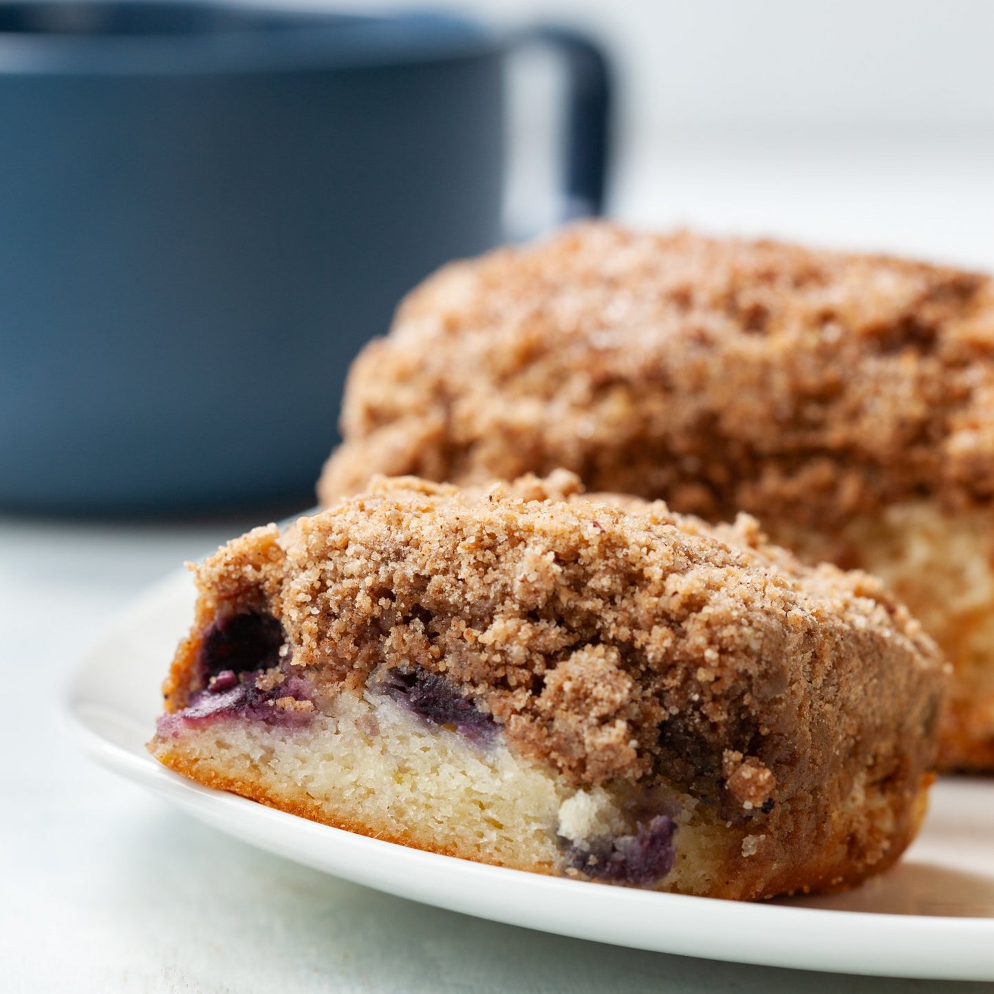 Struesel topped blueberry crumb cake on a hite plate with a blue cup in thee background