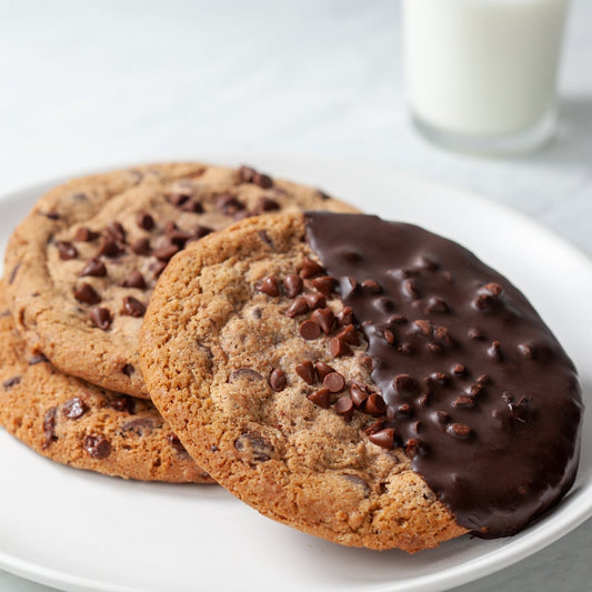 three chcolate chip cookies, one dipped in chocolate on a white plate with a glass of milk in the background,