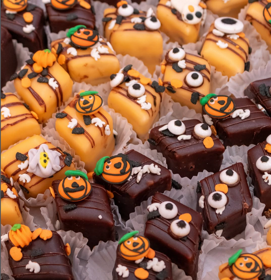 Assorted Halloween-themed petit fours decorated with pumpkins, ghosts, and jack-oh-lanterns in paper pastry cups on a sheet pan.