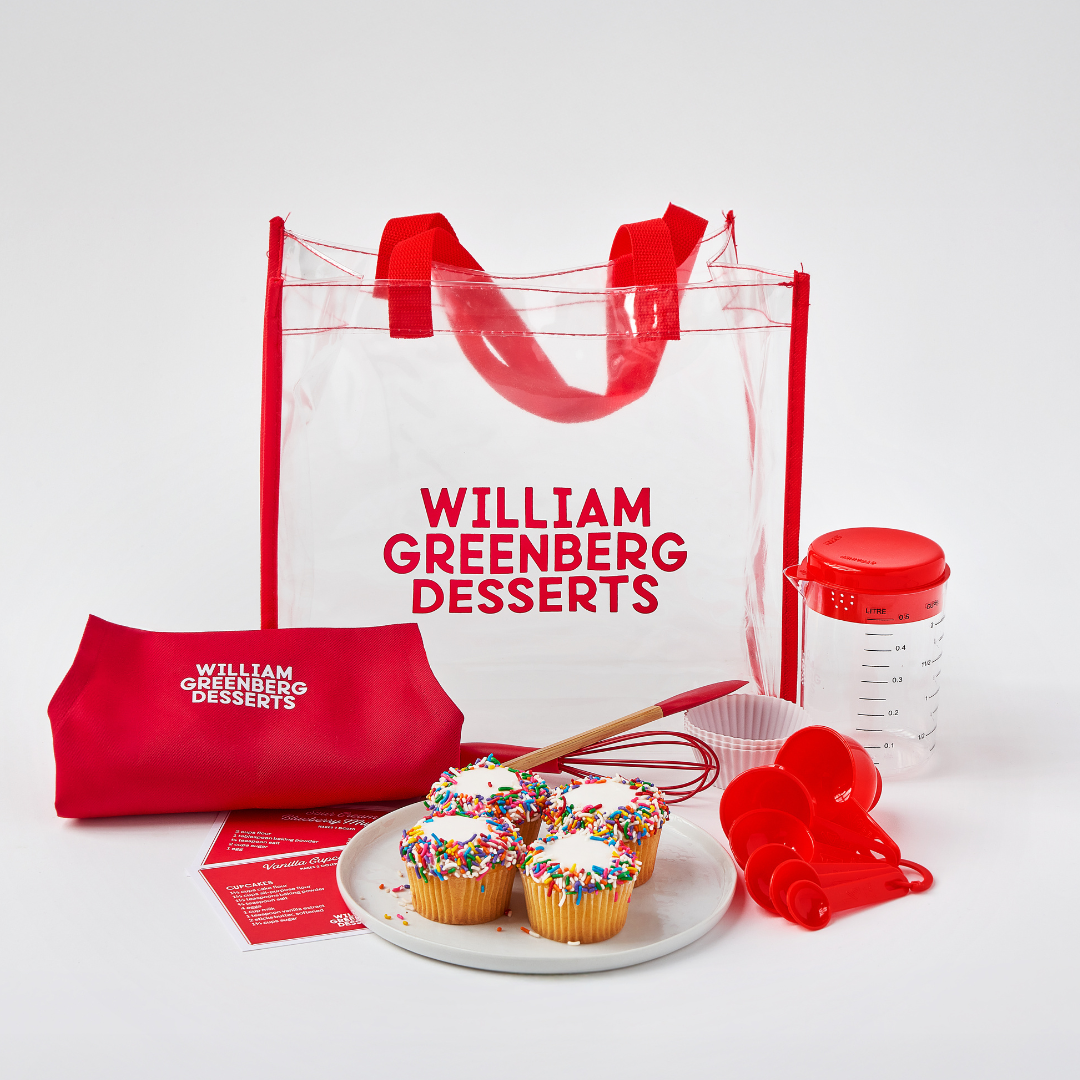 A collection of children's baking tools and ingredients including a red apron, mini kitchen utensils, a measuring cup, silicone baking cups, and a carry bag with the William Greenberg Desserts logo, displayed alongside finished cupcakes.